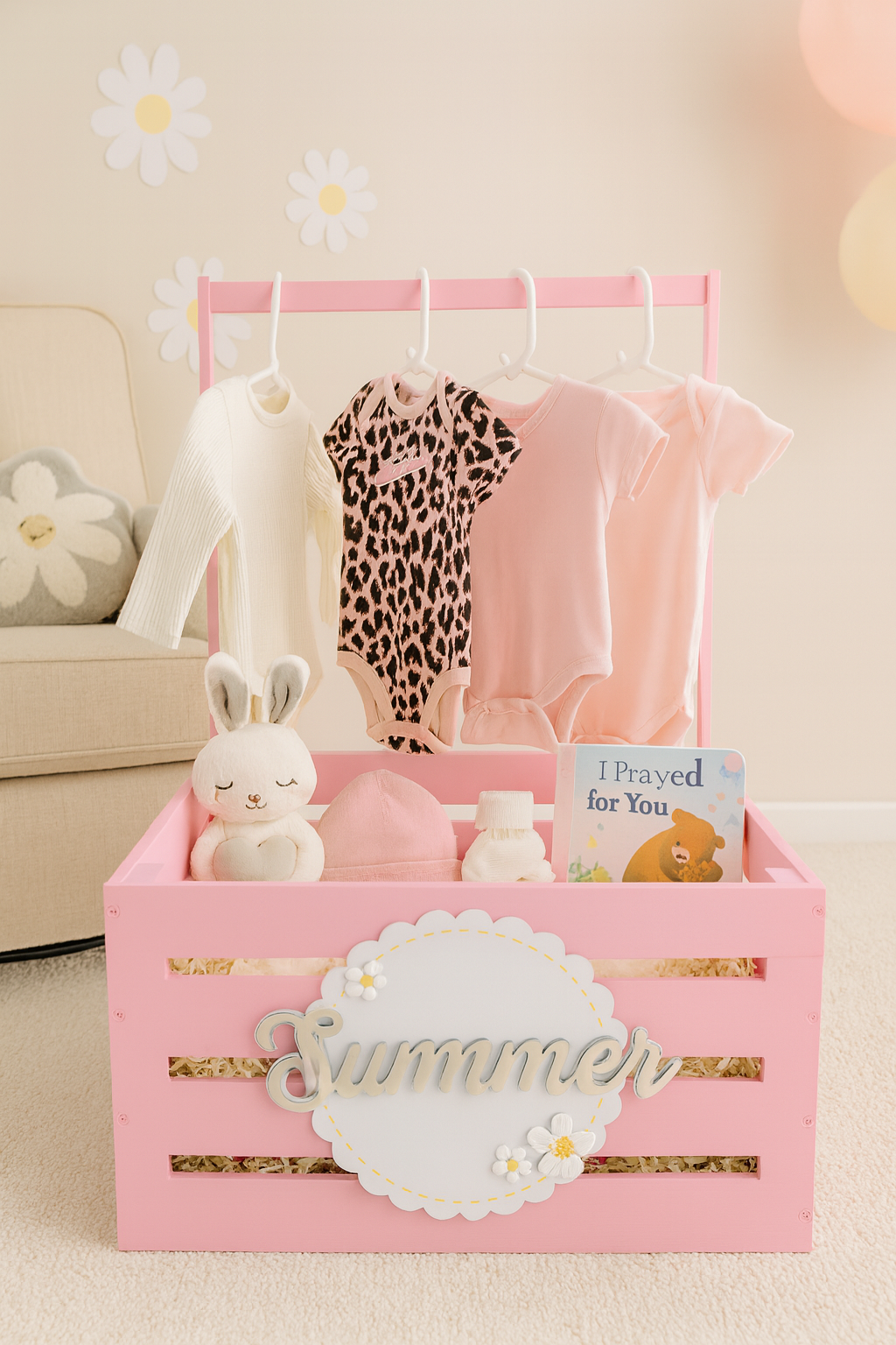 ink personalized baby gift crate labeled “Summer” displayed in a cozy nursery, holding three baby outfits on hangers, a plush bunny toy, baby hat, mittens, socks, and a board book titled “I Prayed for You,” with daisy wall decor and soft pastel balloons in the background.