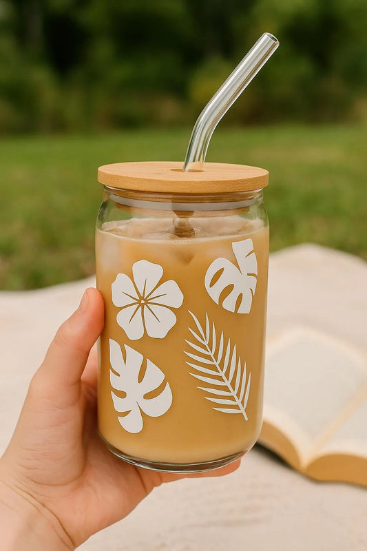 Glass jar with floral design, bamboo lid, and straw held in a hand with a blurred natural background