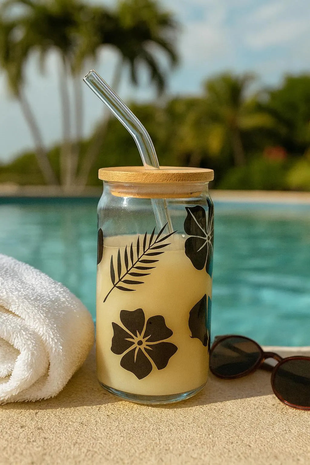 Glass drink container with floral design, straw, and wooden lid by a poolside.