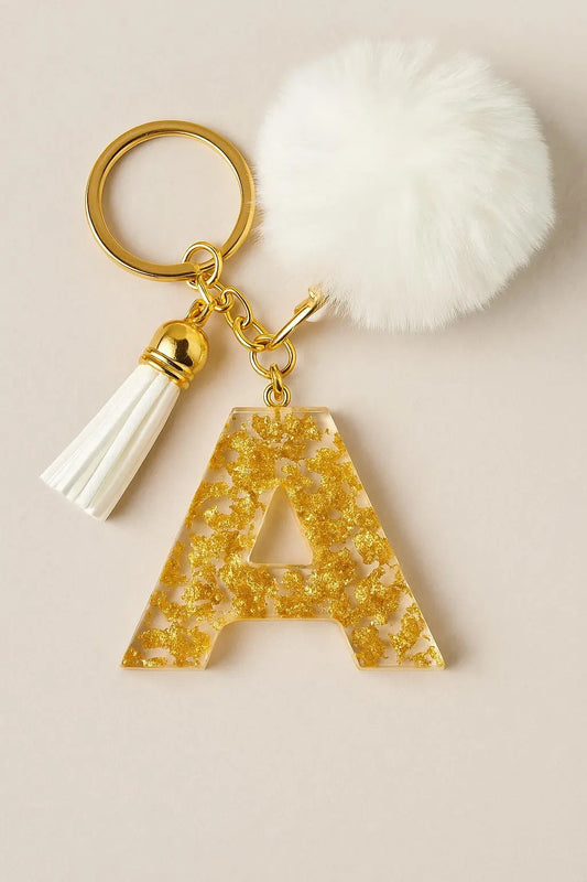 Keychain with gold letter 'A' charm, tassel, and white pom-pom on a beige background