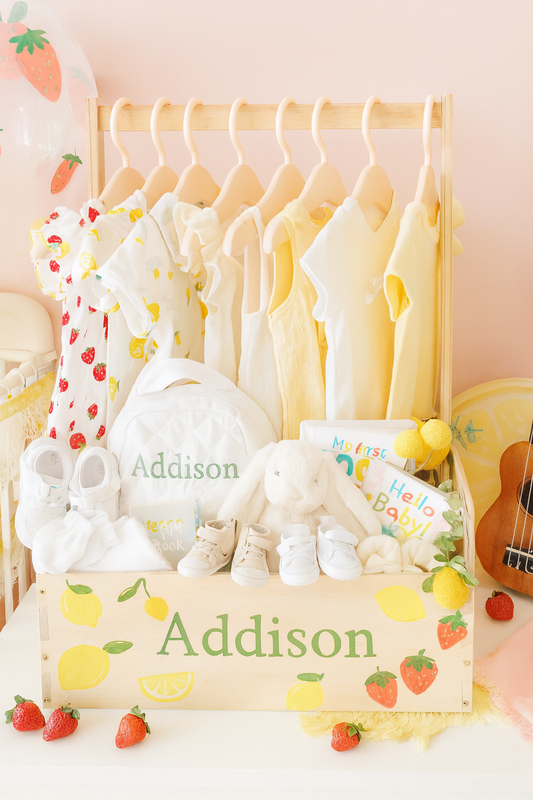 Light wood personalized baby gift crate labeled “Addison” with hand-painted strawberries and lemons, displayed in a nursery, holding six baby outfits in yellow and fruit prints, a white quilted personalized bag, plush bunny, cozy booties, slip-on shoes, and two colorful board books, surrounded by strawberry and lemon decorations.